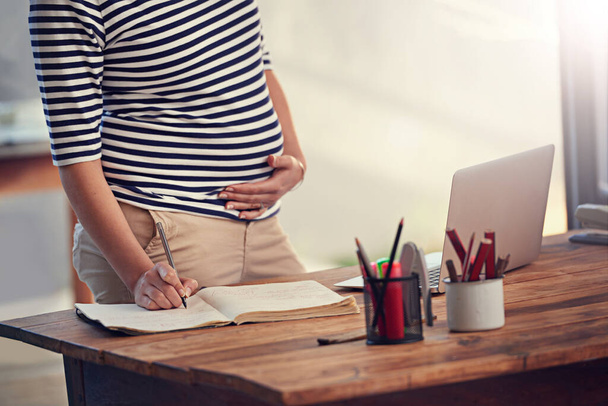 Making sure she knows exactly what needs to be done before maternity leave. Cropped shot of a young pregnant woman writing in her diary while standing in her office. - Фото, изображение