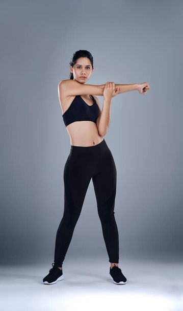 Stretching out before the real workout begins. Full length shot of a sporty young woman standing and stretching her arm against a grey background. - Φωτογραφία, εικόνα