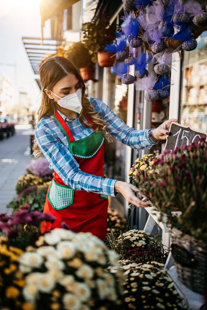Young adult woman working in city street flower shop or florist. She is wearing protective face mask during Covid-19 virus pandemic. Small business concept. - 写真・画像
