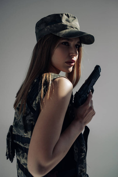 Portrait of young woman in uniform with handgun in her hand against gray background. - Photo, Image