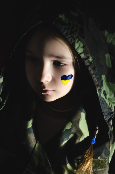 niño molesto con bandera pintada de Ucrania en la mejilla aislado en negro  - Foto, Imagen