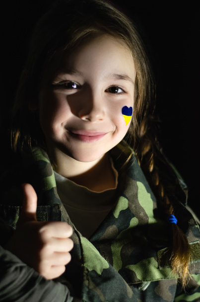 χαμογελαστό παιδί με ζωγραφισμένη ουκρανική σημαία στο μάγουλο που δείχνει σαν απομονωμένο σε μαύρο  - Φωτογραφία, εικόνα