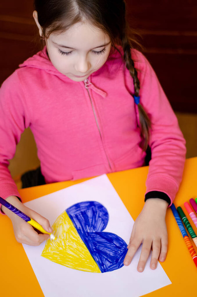 high angle view of girl drawing heart with colors of ukrainian flag on yellow desk  - Zdjęcie, obraz