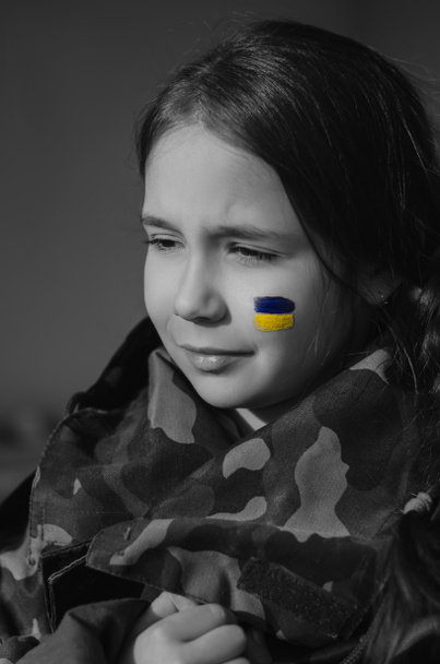Monochromes Foto eines aufgebrachten Kindes mit gemalter ukrainischer Flagge auf der Wange und Tarnjacke - Foto, Bild