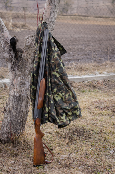 camouflage jacket hanging on tree near rifle in woods - Photo, Image
