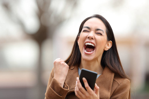 Mujer emocionada sosteniendo teléfono móvil celebrando buenas noticias de pie en un parque - Foto, Imagen
