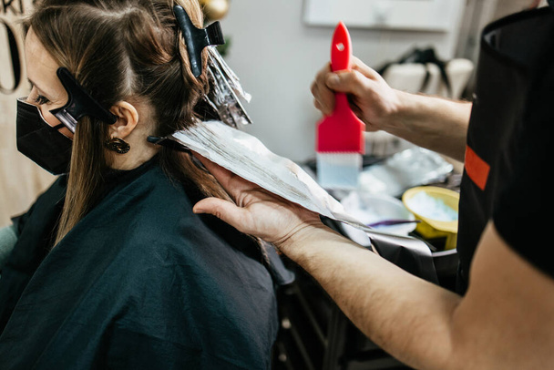 Hairdresser is dyeing female hair, making hair highlights to his client with a foil. She is wearing protective face mask as protection against virus pandemic. - Photo, image