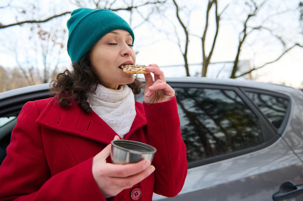 Красивая белая женщина, женщина-водитель, поедающая еду, выносит вкусное печенье и пьет горячий чай из термокружки, стоя возле своей машины в заснеженном лесу. Чудесная зима - Фото, изображение