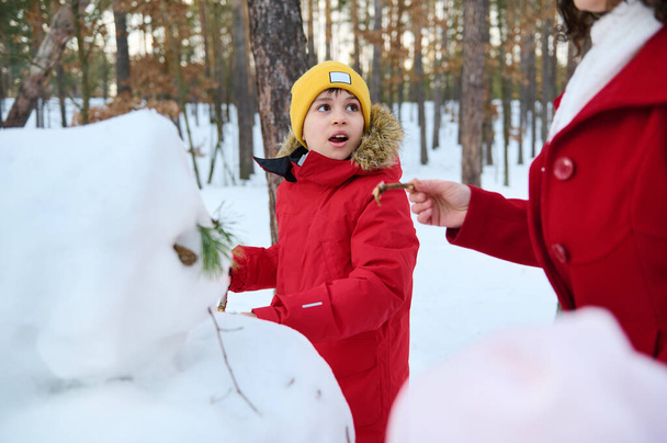 Adorable handsome child, preadolescent Caucasian boy in red down jacket making a snowman, enjoying active weekend pastime with his mom in a snow covered woodland on sunny winter day with cool weather - Photo, Image