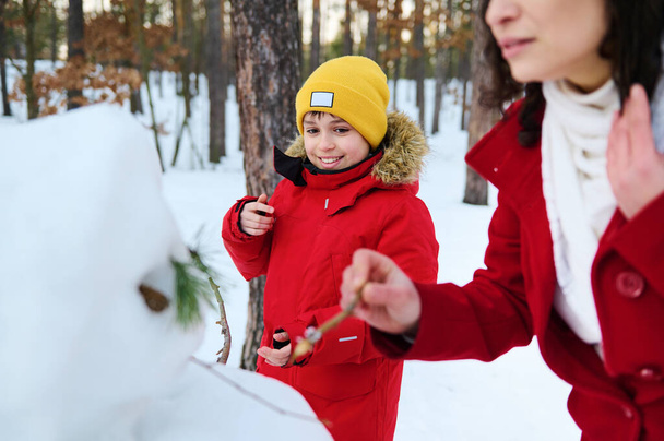 Suloinen komea lapsi, teini-ikäinen eurooppalainen poika punaisessa untuvatakissa tehden lumiukon, nauttien aktiivisesta viikonlopun harrastuksesta äitinsä kanssa lumen peitossa metsässä aurinkoisena talvipäivänä viileällä säällä. - Valokuva, kuva