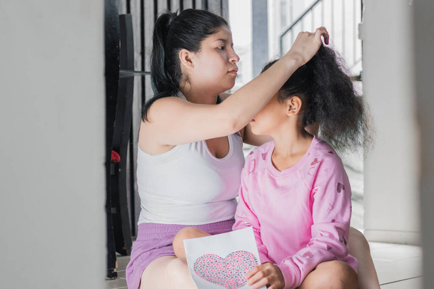 beautiful latina mother with her brunette daughter, combing her hair or making a ponytail while sitting on the floor of her house. young mother talking to her daughter. family concept. - 写真・画像