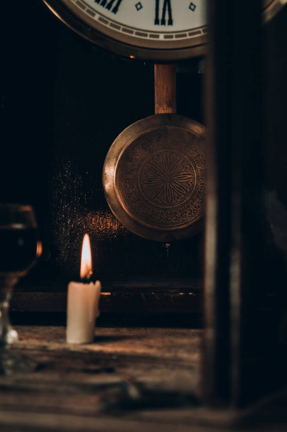 Νεκρή ζωή σε ρουστίκ στυλ με ένα παλιό ρολόι με εκκρεμές, ένα κερί, ένα ποτήρι κονιάκ σε ένα τραχύ ξύλινο τραπέζι - Φωτογραφία, εικόνα
