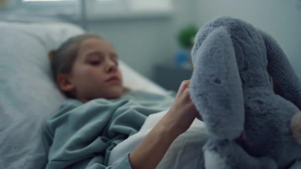 Järkyttynyt tyttö makaa sairaalan sängyssä muotokuva. Surullinen lapsi leikkii pehmolelu yksin. - Materiaali, video