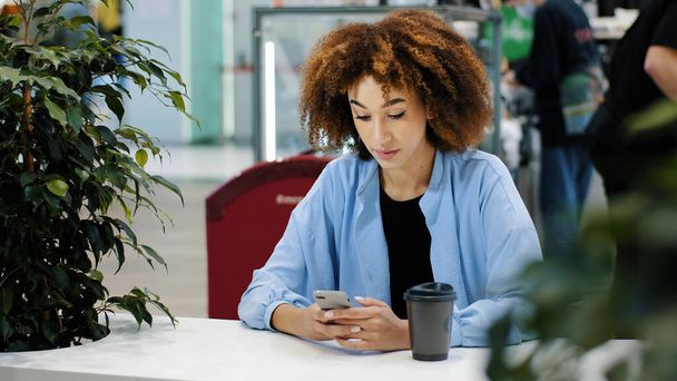 African 20s woman multiracial biracial lady with curly haircut sitting at cafe table drinking coffee tea from paper cup looking in mobile phone typing smartphone chatting browsing buying online app - Photo, Image