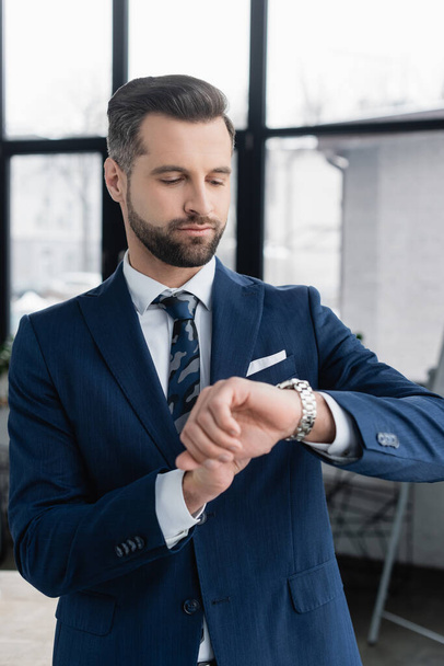 businessman in blazer looking at wristwatch in blurred office - Fotoğraf, Görsel