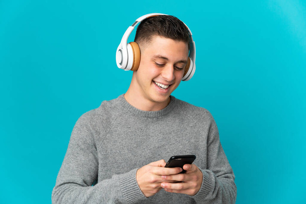 Jovem caucasiano isolado no fundo azul ouvindo música e olhando para celular - Foto, Imagem