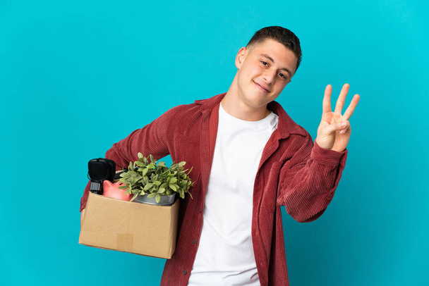 Young caucasian man making a move while picking up a box full of things isolated on blue background happy and counting three with fingers - Fotó, kép