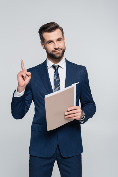 positive businessman holding folder and showing idea gesture isolated on grey - Zdjęcie, obraz