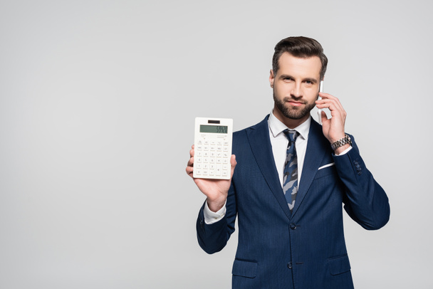 economista en traje azul mostrando calculadora y hablando en el teléfono móvil aislado en gris - Foto, imagen
