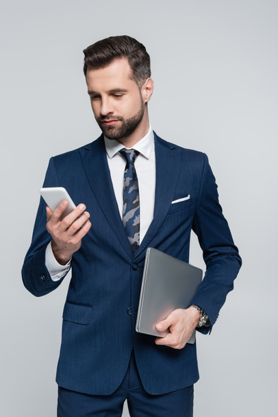 brunette businessman with laptop looking at mobile phone isolated on grey - Φωτογραφία, εικόνα