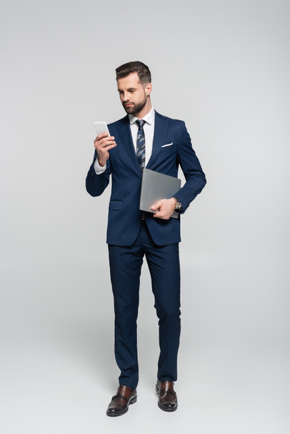 full length view of businessman with laptop using cellphone isolated on grey - Foto, afbeelding