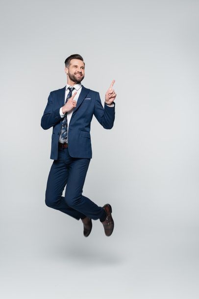 cheerful businessman levitating and pointing with fingers on grey - Foto, Imagen
