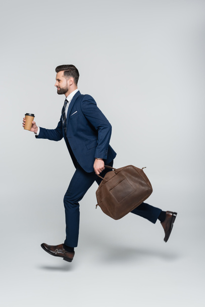 vista lateral del hombre de negocios en traje azul corriendo con maletín y taza de papel en gris - Foto, Imagen