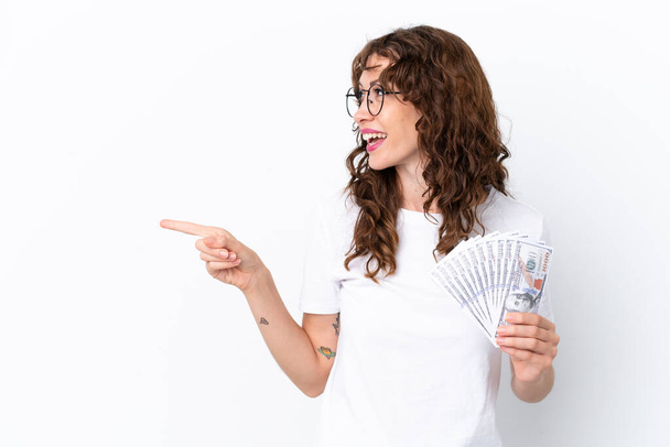 Young woman with curly hair taking a lot of money isolated background on white background pointing finger to the side and presenting a product - Foto, afbeelding