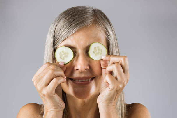 Mature woman with cucumber slices on eyes in studio - Fotografie, Obrázek