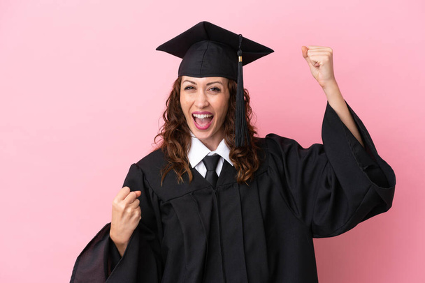 Fiatal egyetemi diplomás nő elszigetelt rózsaszín háttér ünnepli a győzelem - Fotó, kép