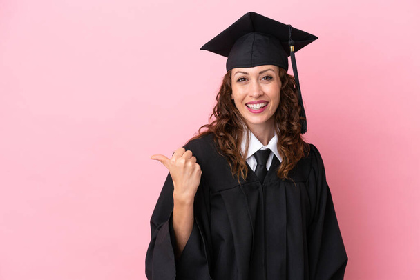 Giovane laureata donna isolata su sfondo rosa che punta verso il lato per presentare un prodotto - Foto, immagini