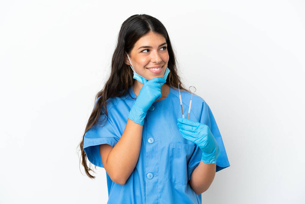 Mujer dentista sosteniendo herramientas sobre un fondo blanco aislado mirando hacia un lado y sonriendo - Foto, Imagen