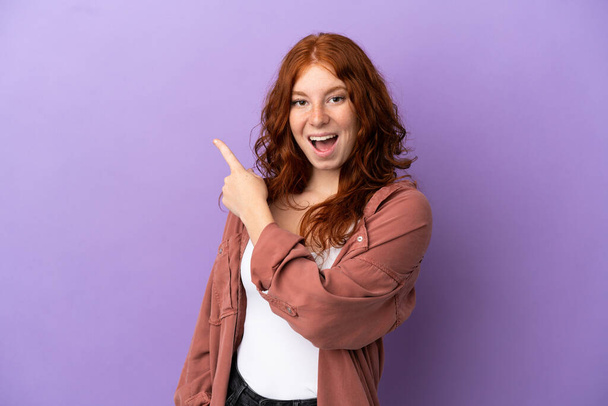 tiener roodharige meisje over geïsoleerde paarse achtergrond verrast en wijzen kant - Foto, afbeelding
