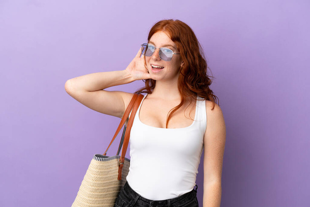 Teenager redhead girl holding a beach bag isolated on purple background listening to something by putting hand on the ear - Foto, afbeelding