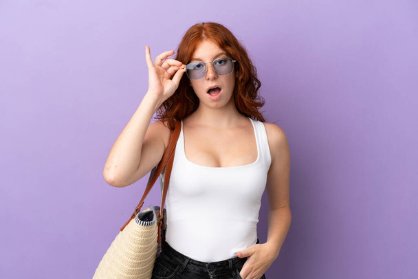 Teenager redhead girl holding a beach bag isolated on purple background with glasses and surprised - Fotografie, Obrázek