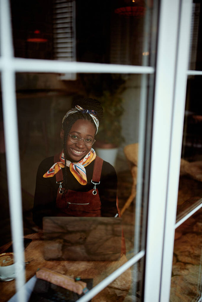 A smart African college girl sits in the cafeteria and studies for an exam. The picture was taken from the outside. A girl on student exchange sitting in a coffee shop and studying. - Zdjęcie, obraz