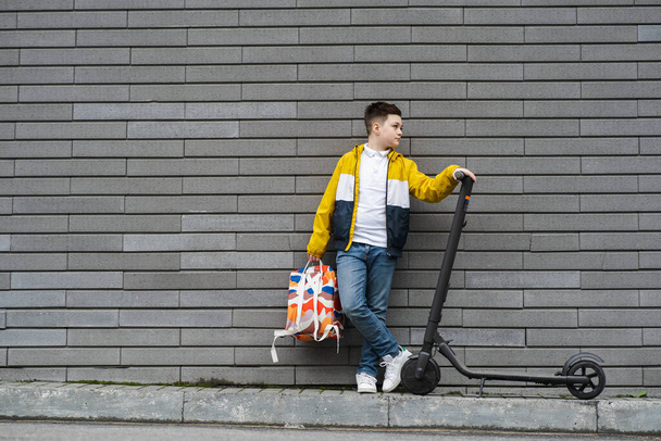 Nowoczesny nastolatek z plecakiem i skuterem elektrycznym stoi przy ceglanej ścianie - Zdjęcie, obraz