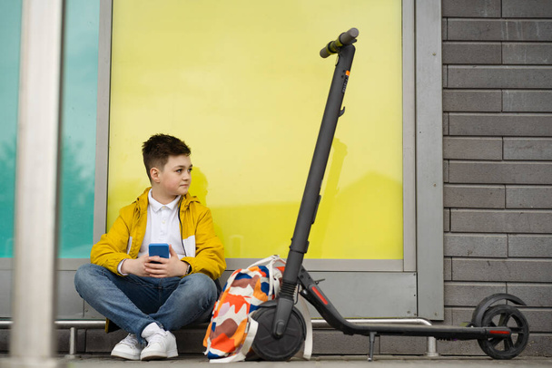 Knappe tiener zit op straat naast zijn scooter en rugzak - Foto, afbeelding