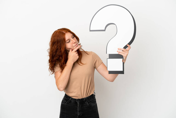 Tini vörös hajú lány elszigetelt fehér háttér kezében egy kérdőjel ikon és kétségei vannak - Fotó, kép