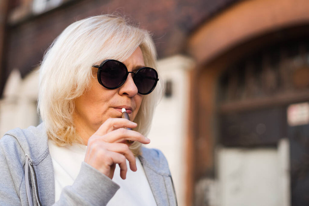 Middle aged blonde European woman smoking heated tabacco e-cigarette outdoors on the street. Alternative smoking way - Fotografie, Obrázek