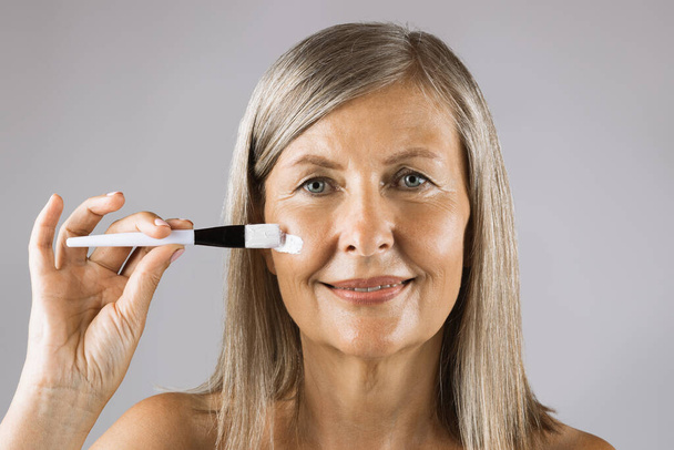 Attractive aged lady using cosmetic brush for applying cream - Foto, afbeelding