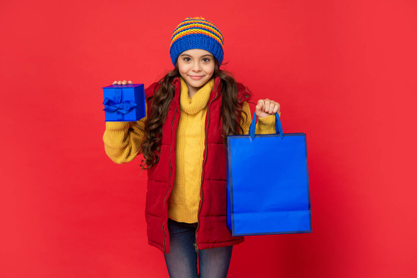 cheerful teen girl wearing knitted hat hold shopping bag and present box on red background, shopping - 写真・画像
