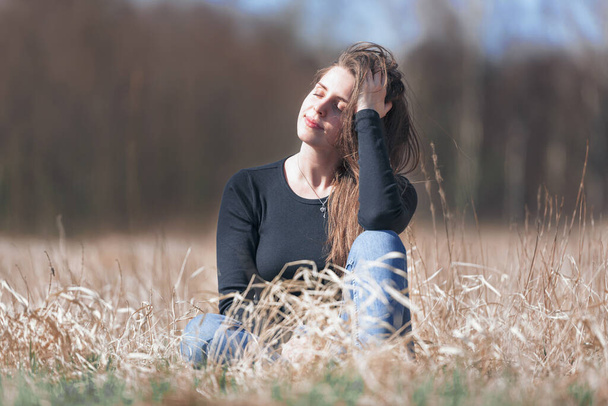Egy alkalmi ruhás fiatal hölgy ül a fűben. A lány élvezi a napsugarakat, amik ráhullanak. A fű száraz, és erdő van a háttérben. Ez egy napos nap.. - Fotó, kép