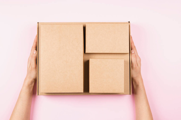 Horní pohled na hromadu lepenkových krabic v ženských rukou. Prázdné hnědé balíčky krabice na pastelově růžové pozadí. Balení, nakupování, doprava zdarma, dodací koncepce - Fotografie, Obrázek