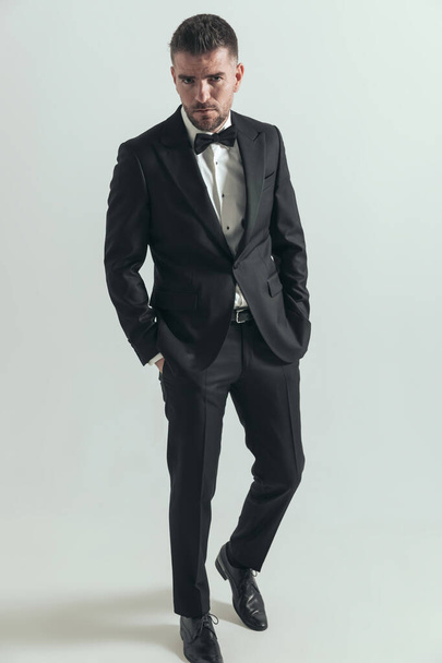 hombre de negocios guapo posando con una buena postura, tomados de la mano en bolsillos sobre fondo gris - Foto, Imagen