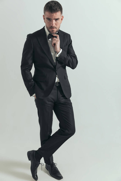 cool sexy fashion model crossing his feet, holding a hand in pocket and arranging his bowtie on gray background - Foto, Bild
