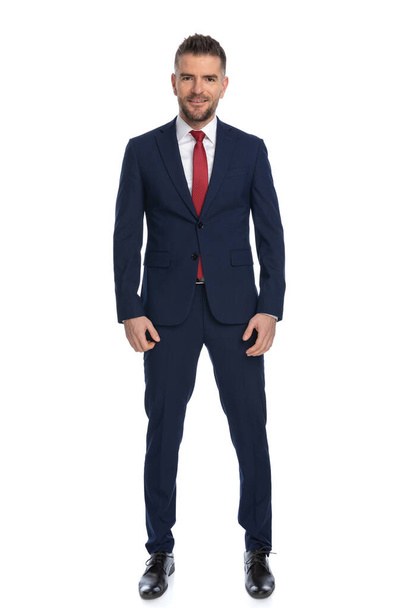 elegant guy dressed with a navy suit with red tie and posing with a smile on his face against white background - Foto, Imagen