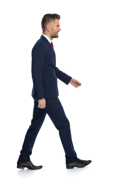 vista lateral de un hombre de negocios vistiendo un traje azul marino y caminando en su camino contra el fondo blanco - Foto, Imagen