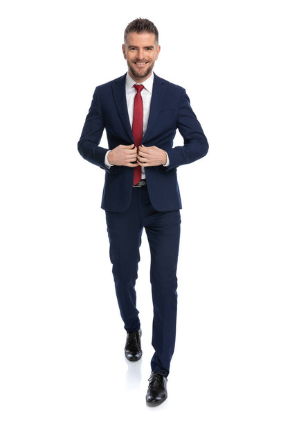 joven hombre de negocios desabrochándose la chaqueta, sonriendo y caminando hacia la cámara sobre fondo blanco - Foto, imagen
