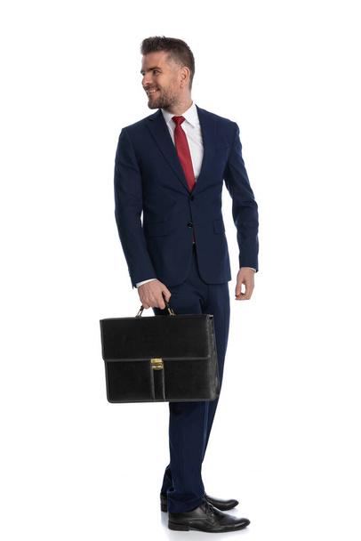 hombre de negocios guapo mirando hacia atrás por encima del hombro y sonriendo mientras sostiene su caso - Foto, imagen